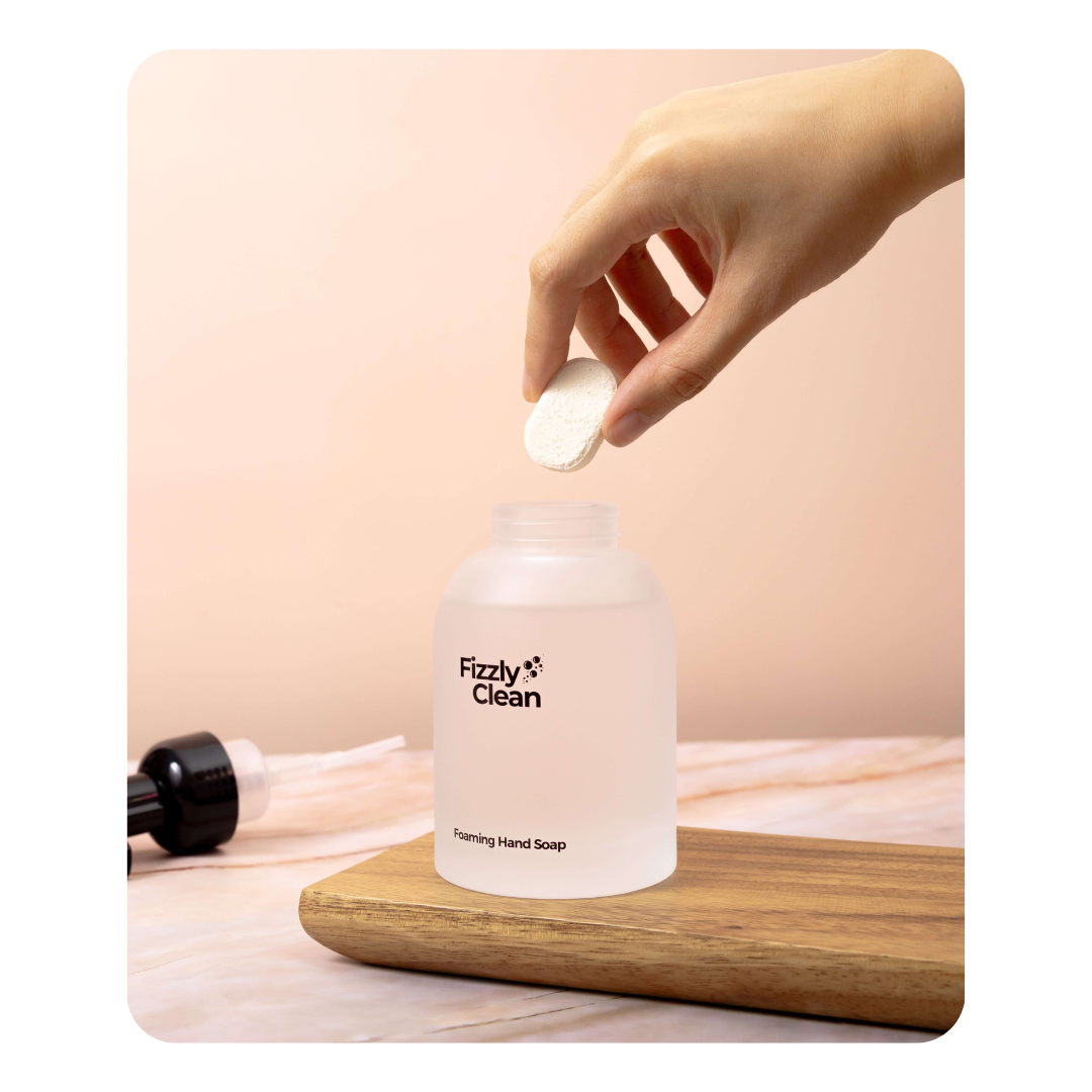Hand holding a bar of soap above a bottle labeled 'Fizzy Clean' on a wooden surface with a pink background.